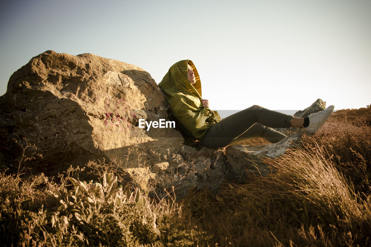 Side view of man with blanket sitting on rock against clear sky