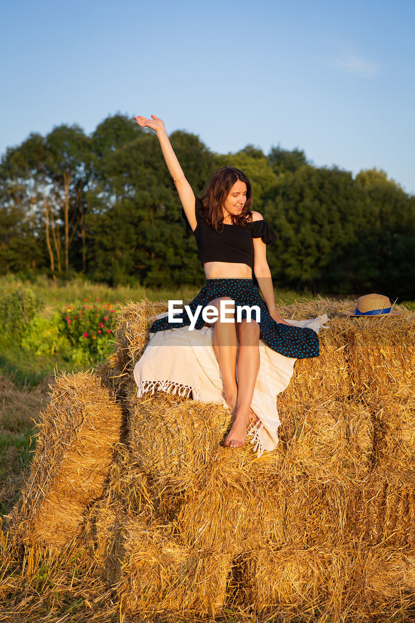 Woman looking at the setting sun and waving her hand, sitting on a large pile of straw bales