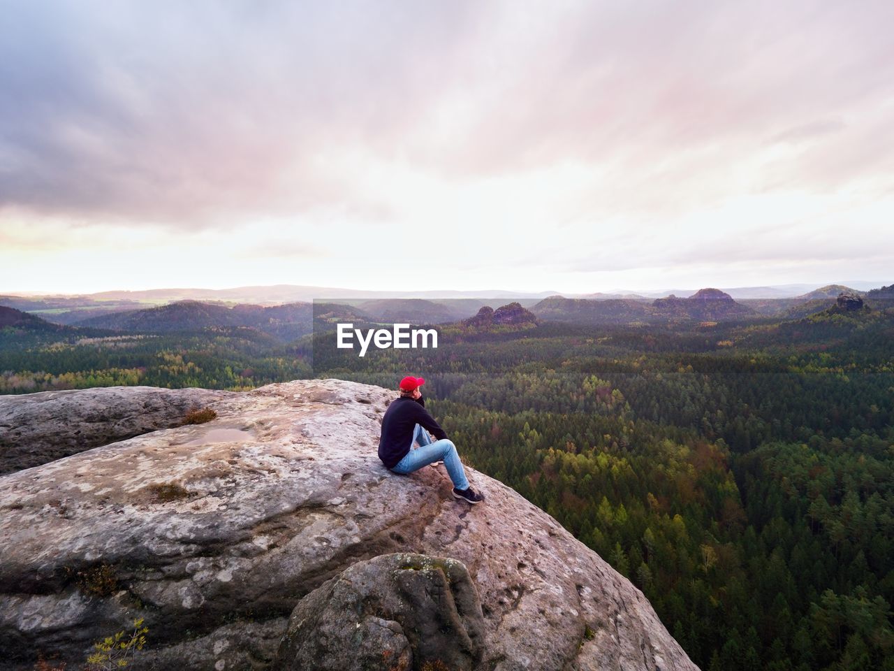 Hiker man on cliff. tourist in red cap, black sweatshirt and jeans.. cold morning sun hiidden 