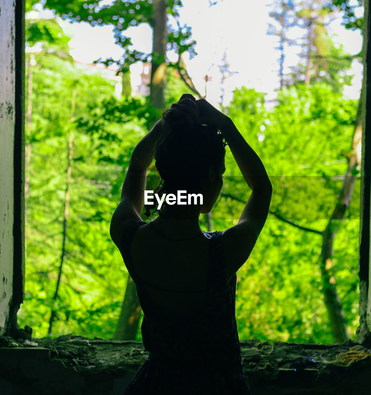 Rear view of woman standing at abandoned window against trees