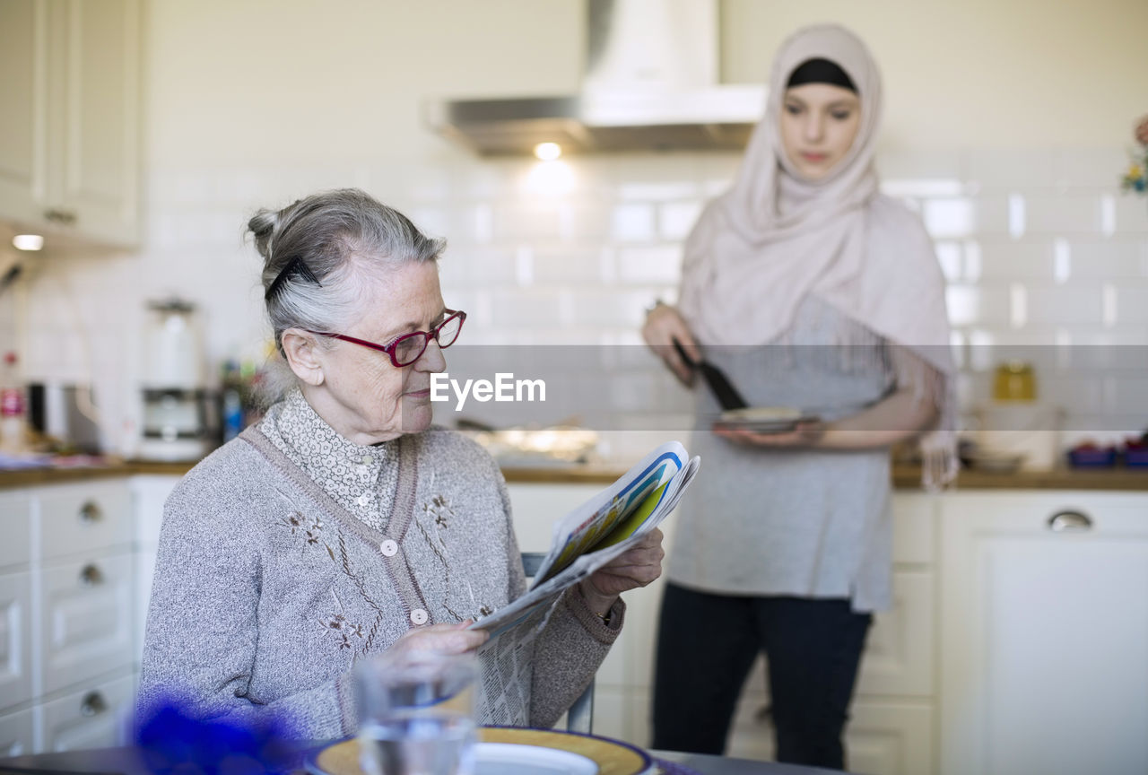 Senior woman reading newspaper with female home caregiver preparing food in kitchen