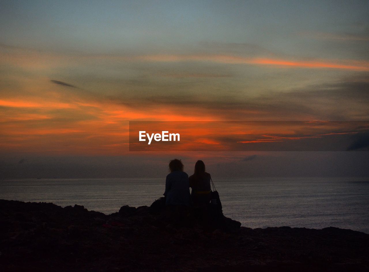 Rear view of couple sitting on coast against cloudy sky at dusk
