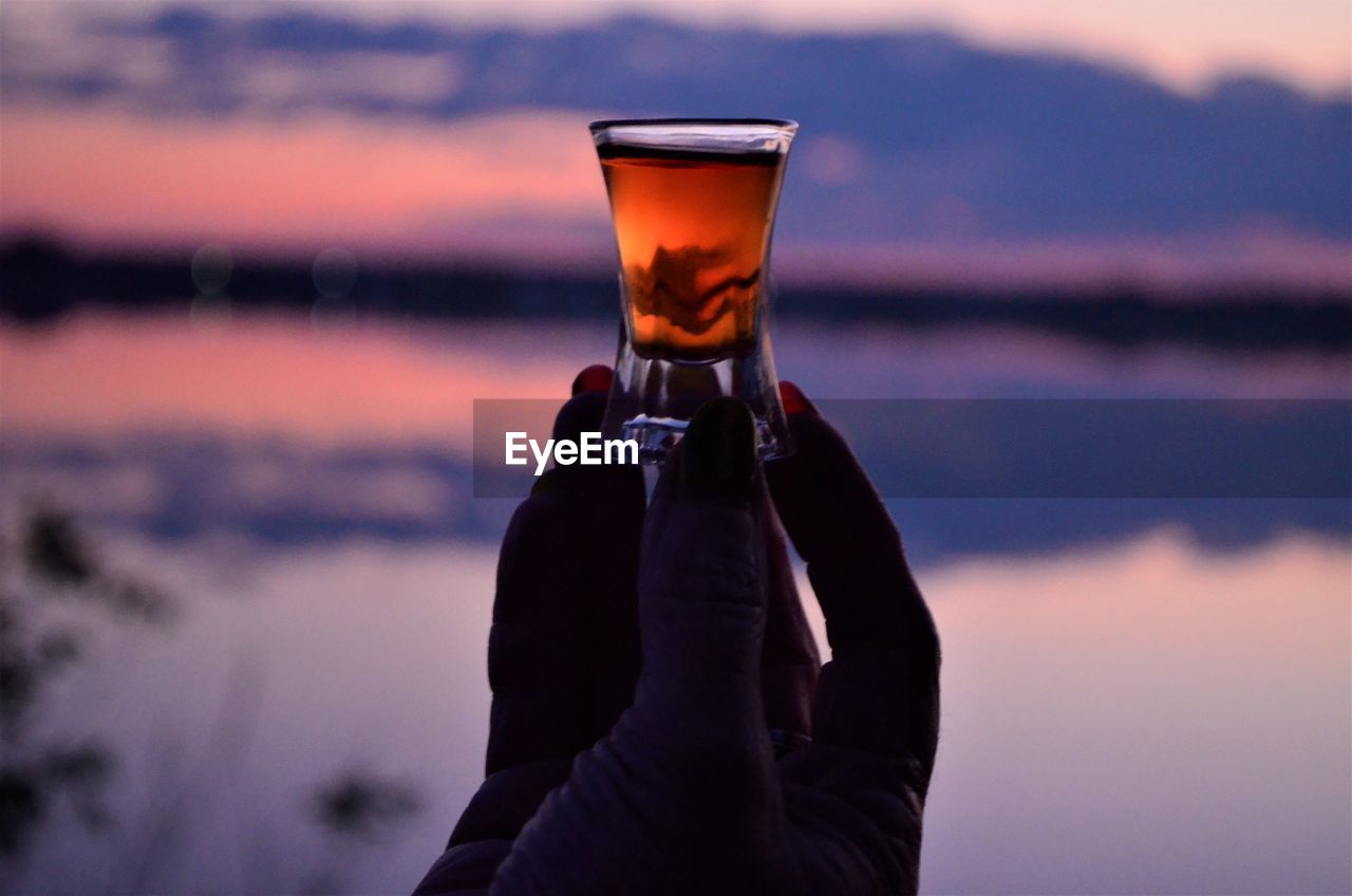 Added a female hand holding a cognac glass against the sky during sunset on the river