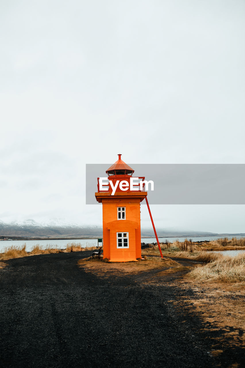 Orange lighthouse on coast between wild lands near water surface and cloudy sky