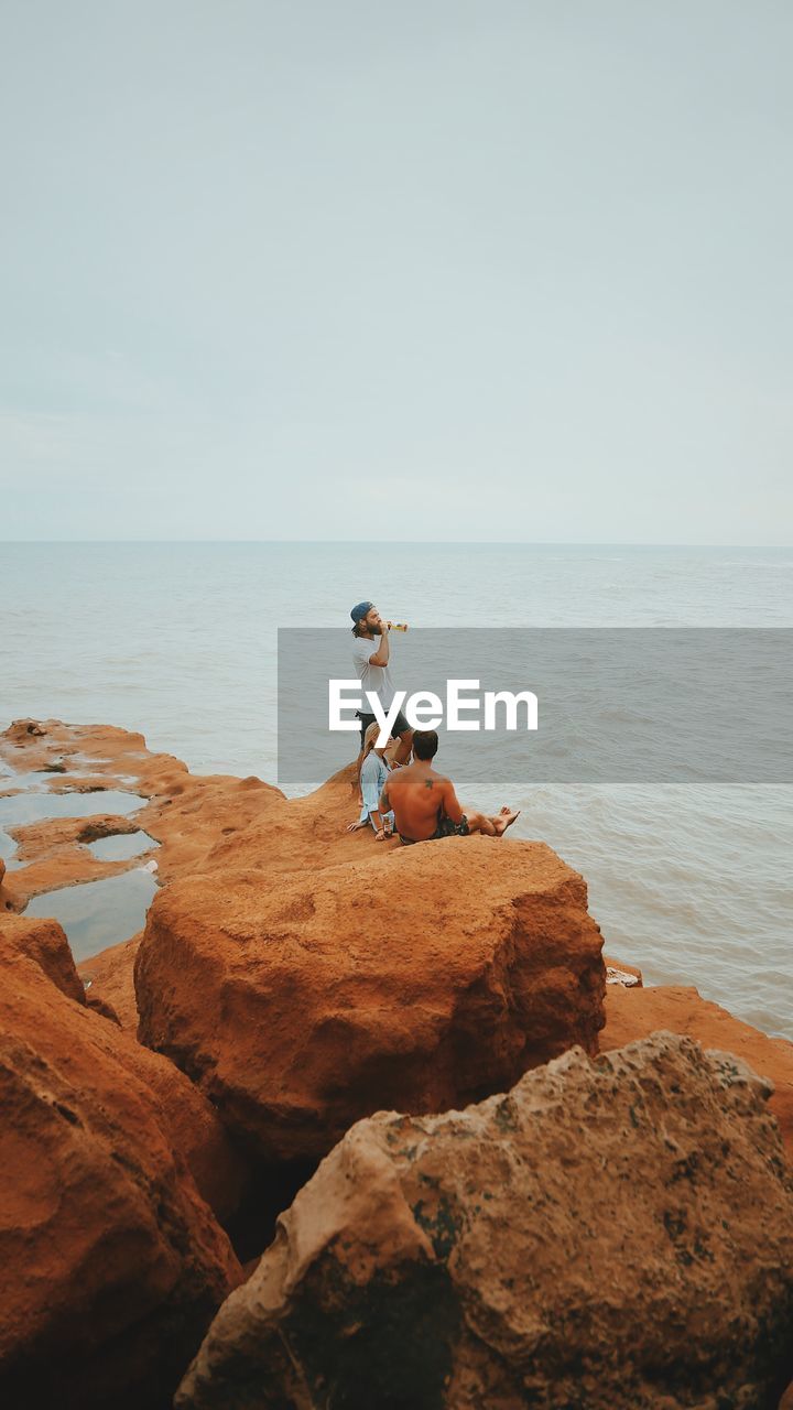 MAN STANDING ON ROCK AT BEACH AGAINST SKY