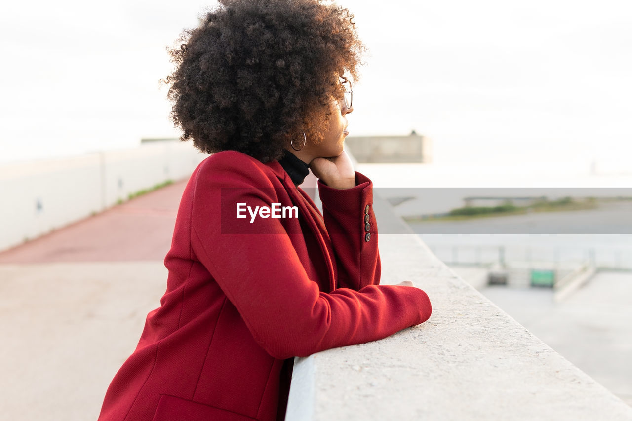 Side view of african american female with afro hairstyle wearing jacket leaning on barrier of embankment of river and looking away