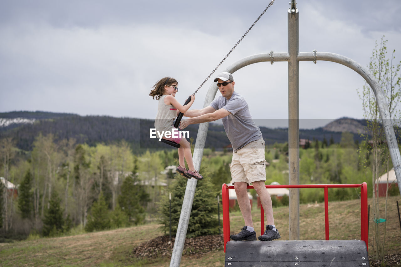 Father pushing daughter sitting on chain swing against cloudy sky at park