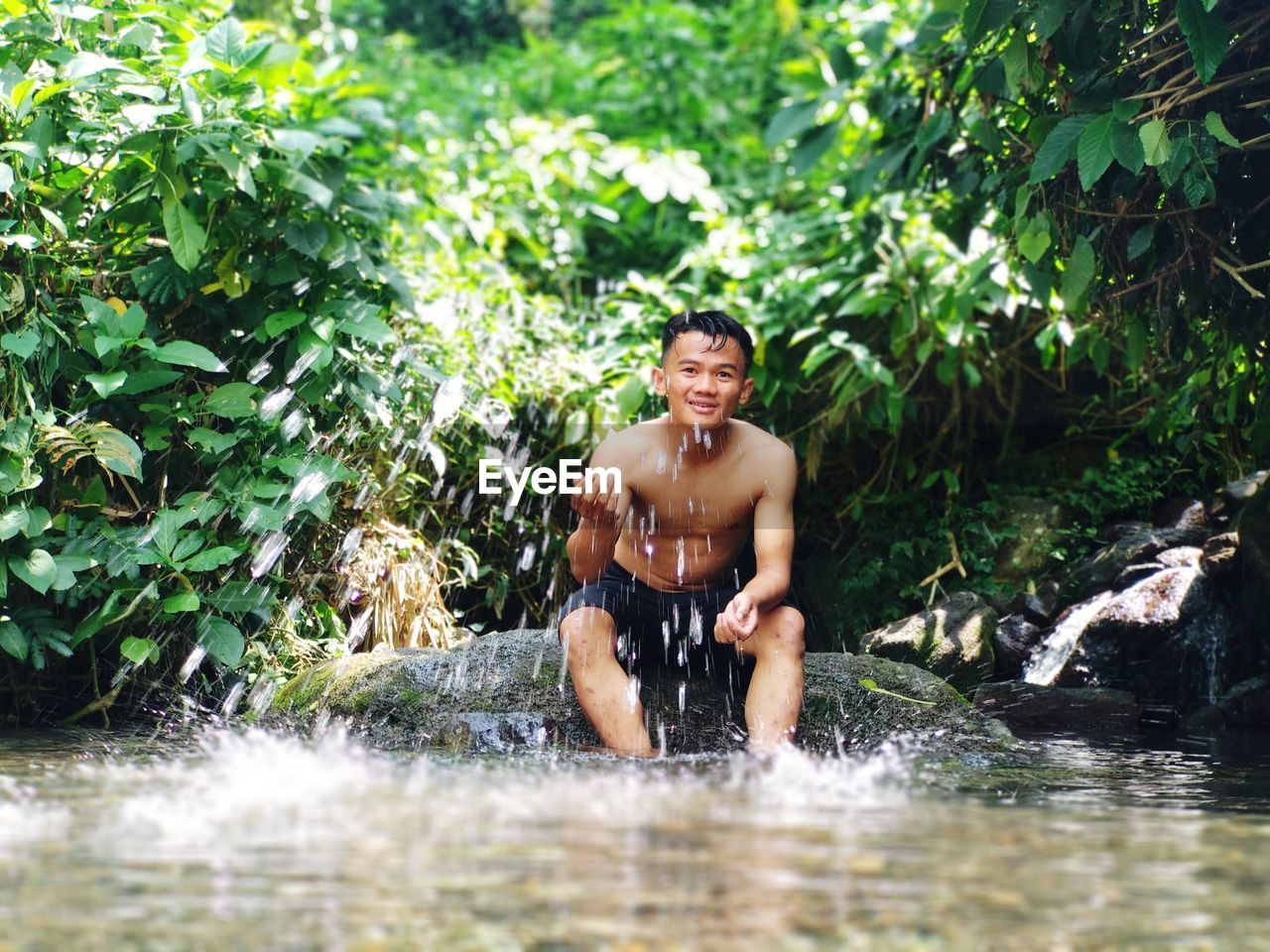 Portrait of shirtless man smiling while sitting on rock by lake