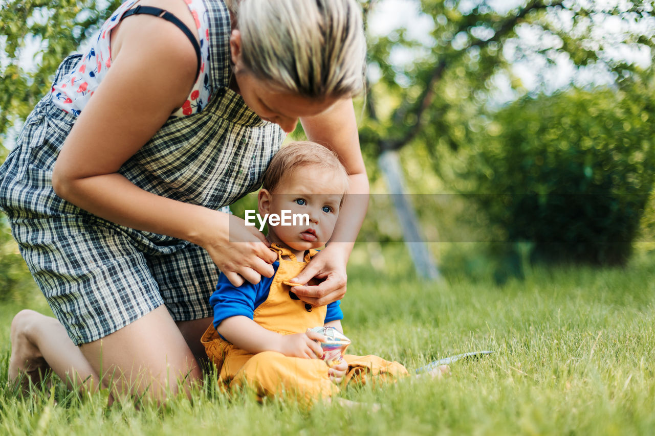 A young mother sits with her young son on the grass in the garden and adjusts his clothes.