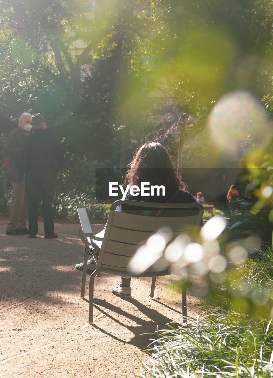 REAR VIEW OF PEOPLE SITTING ON BENCH AT PARK