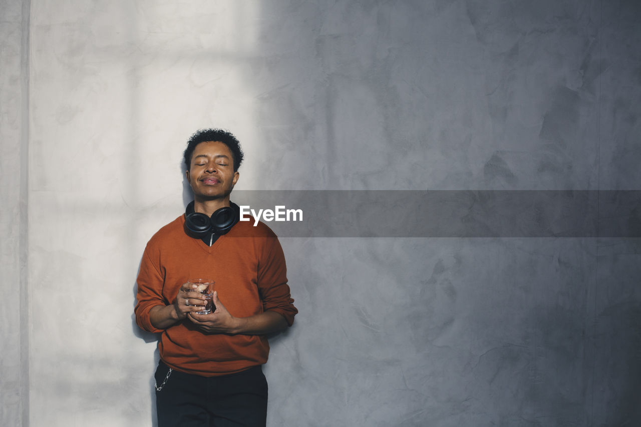 Male entrepreneur holding glass with eyes closed standing against wall