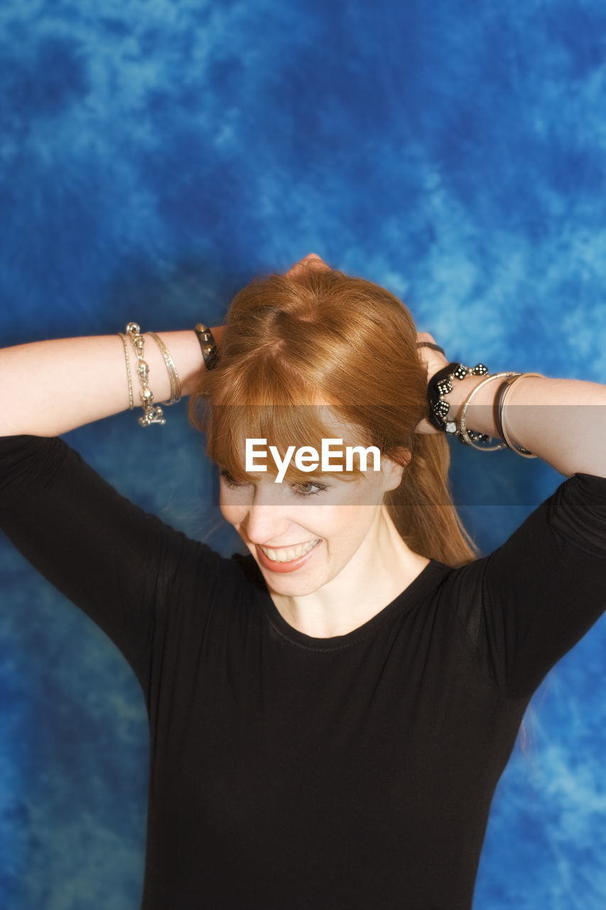 Smiling young woman tying hair while standing against blue background