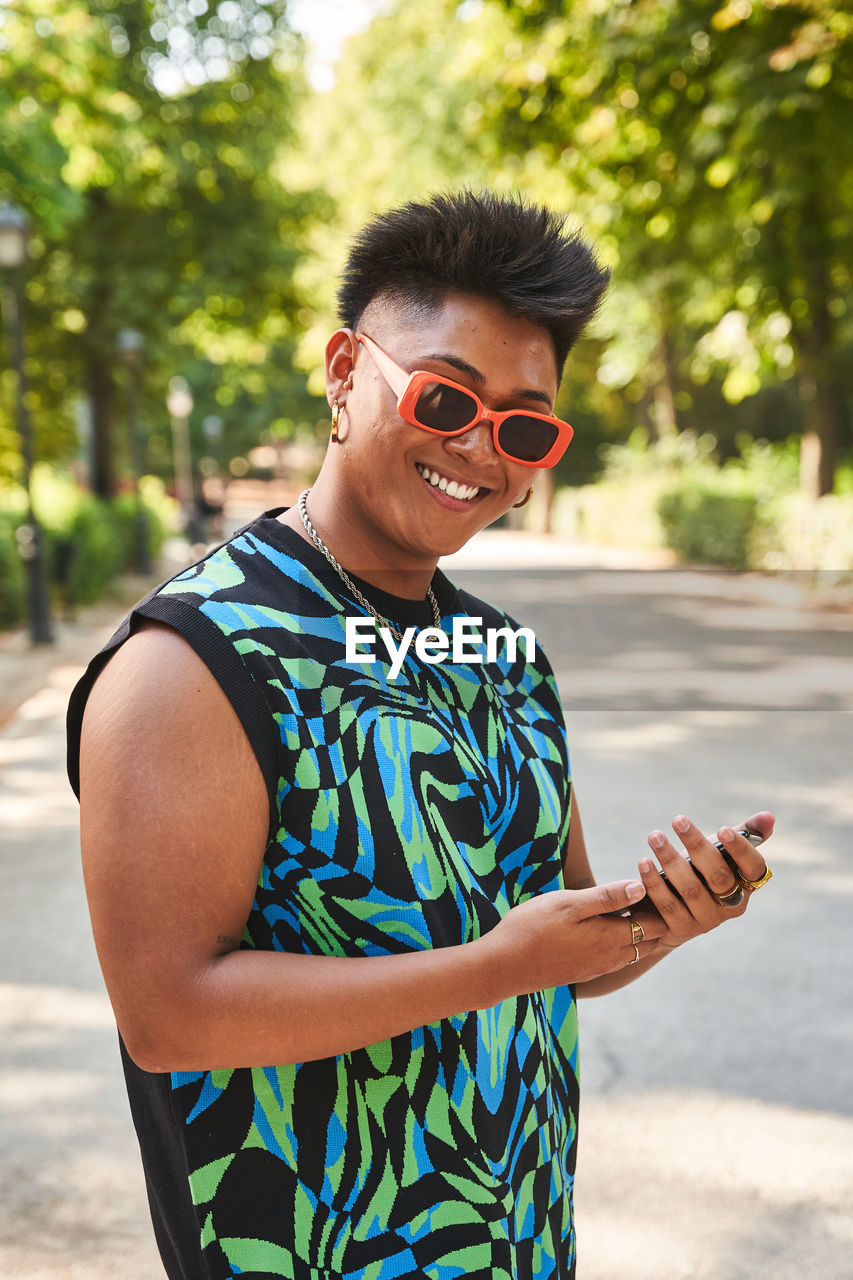 Happy filipino transgender woman in stylish clothes and sunglasses browsing cellphone and looking at camera with smile while standing on sunlit path in summer in park