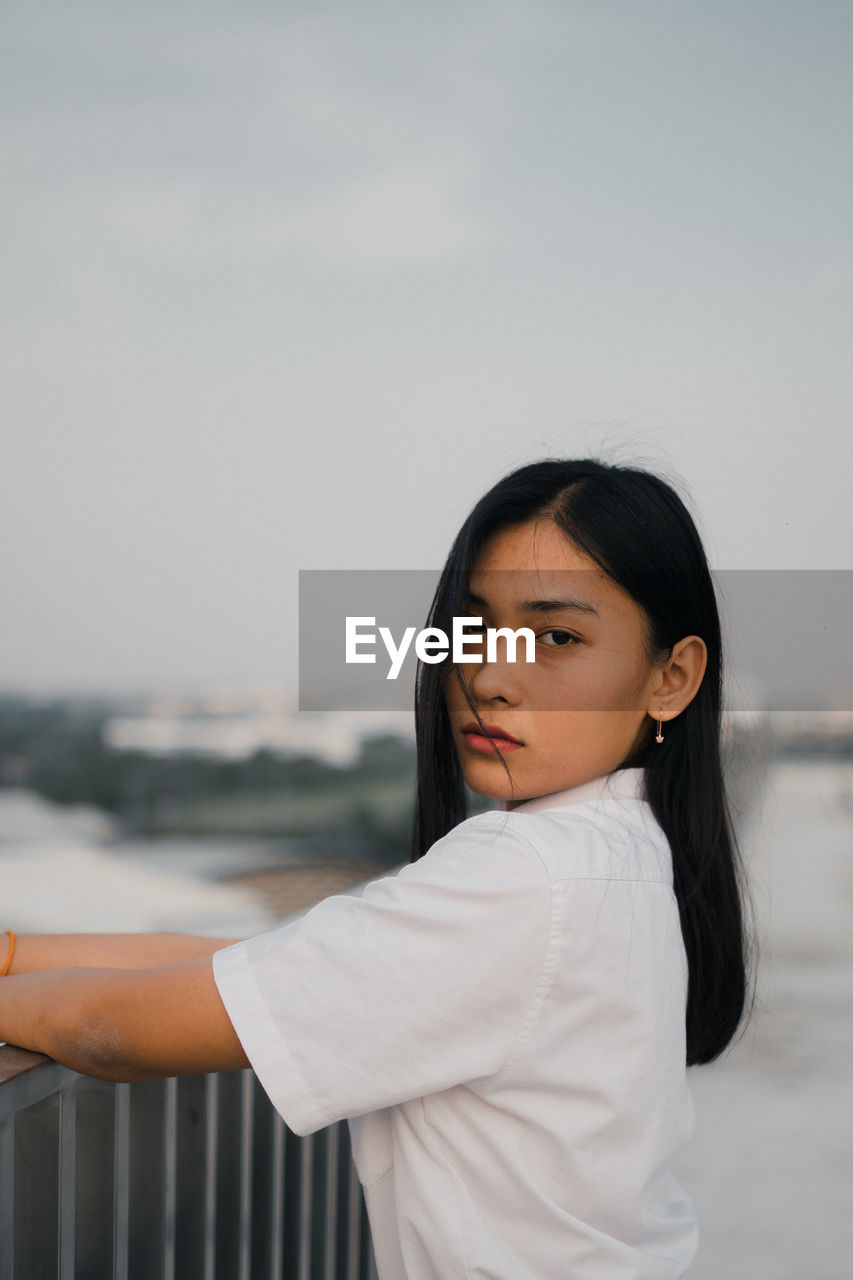 Portrait of young woman standing by railing against sky