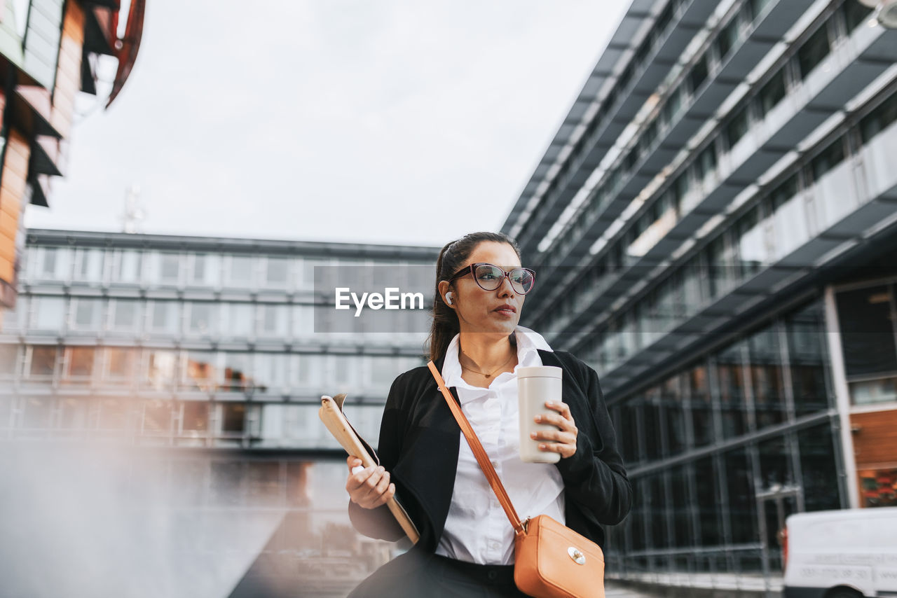 Businesswoman looking away holding insulated drink container while standing in front of building