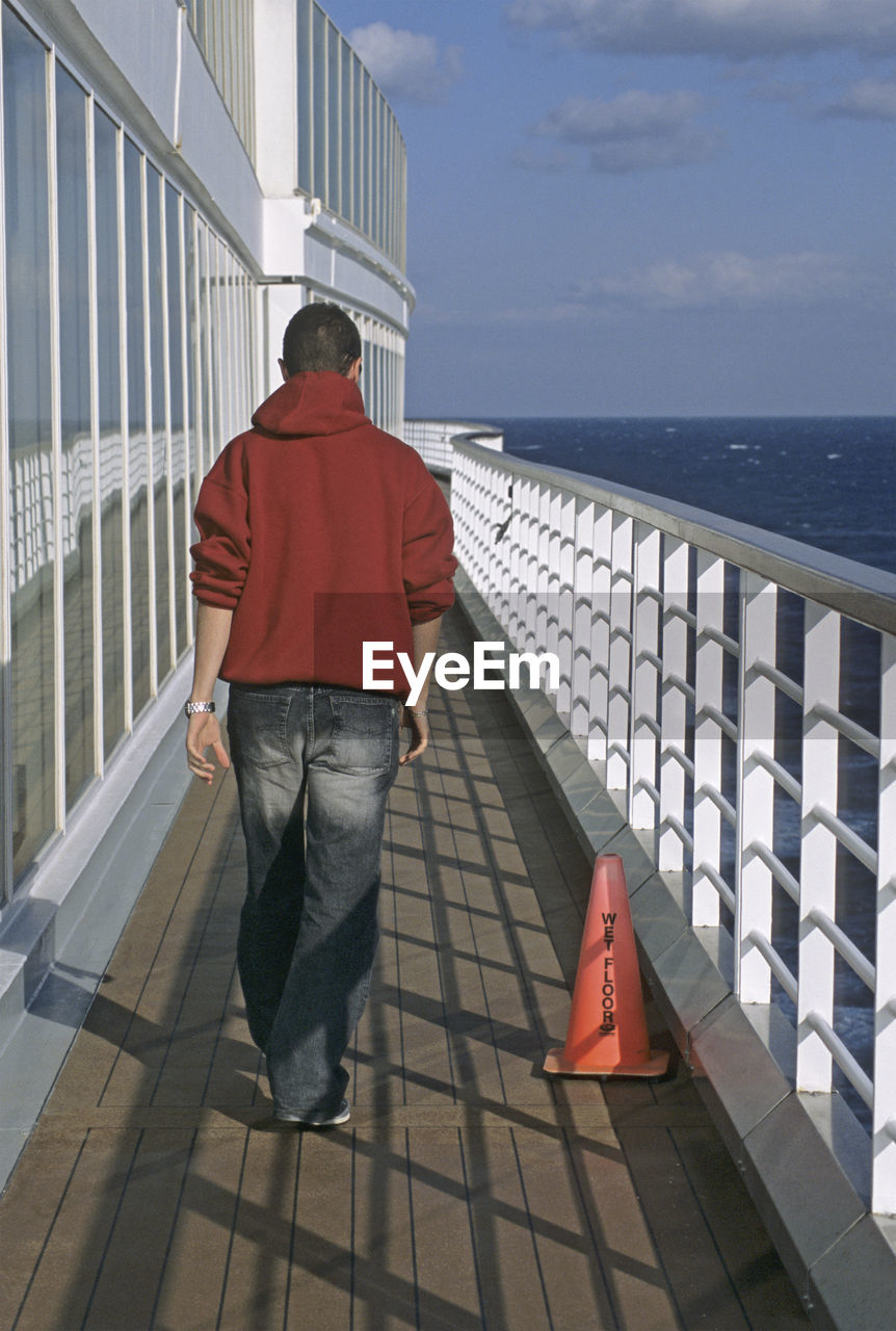 REAR VIEW OF MAN WALKING ON RAILING BY SEA AGAINST SKY