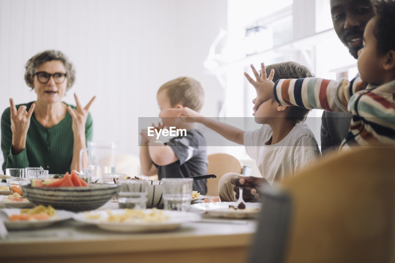 Preschool students gesturing at teacher while sitting at dining table during lunch break