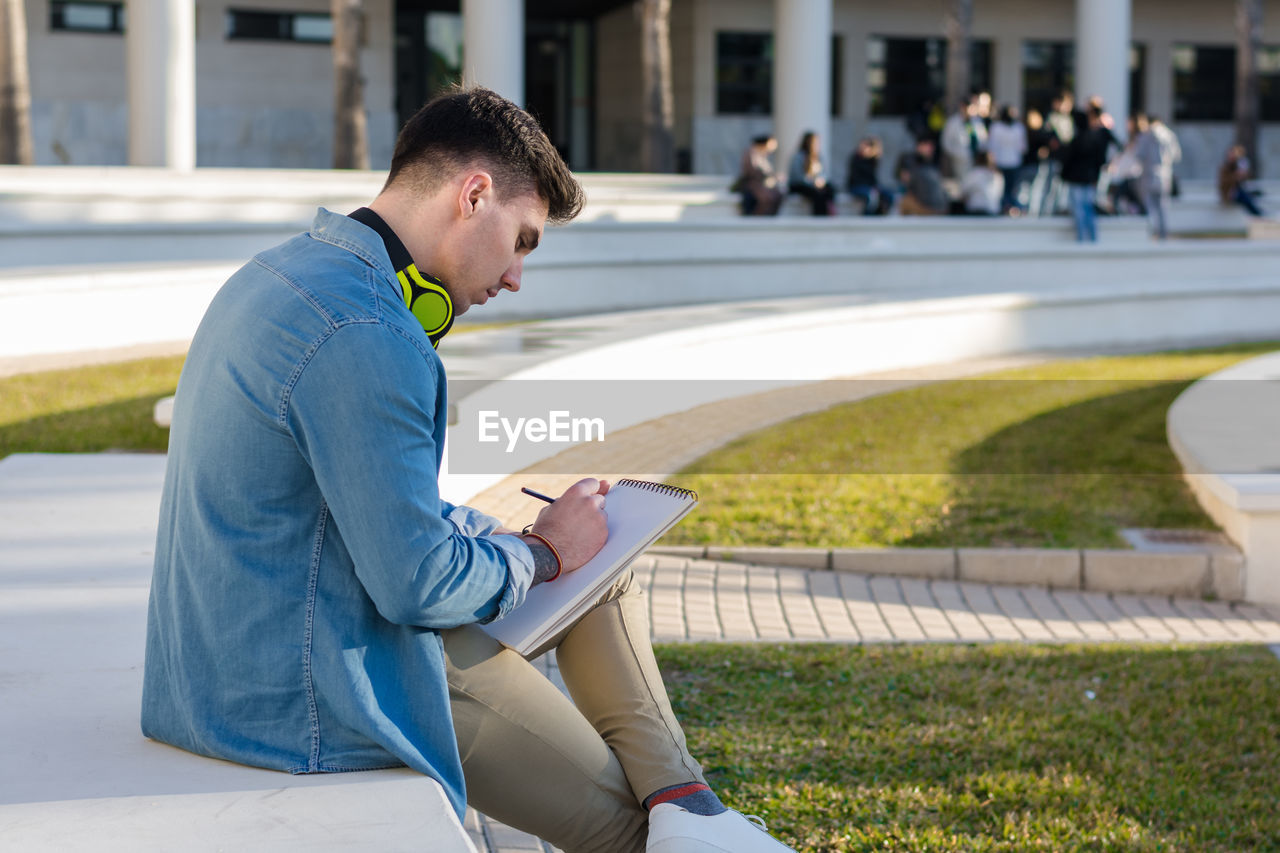 Side view of thoughtful smart male student in bright headphones studying in university square and writing in note book sitting on fence with crossed leg