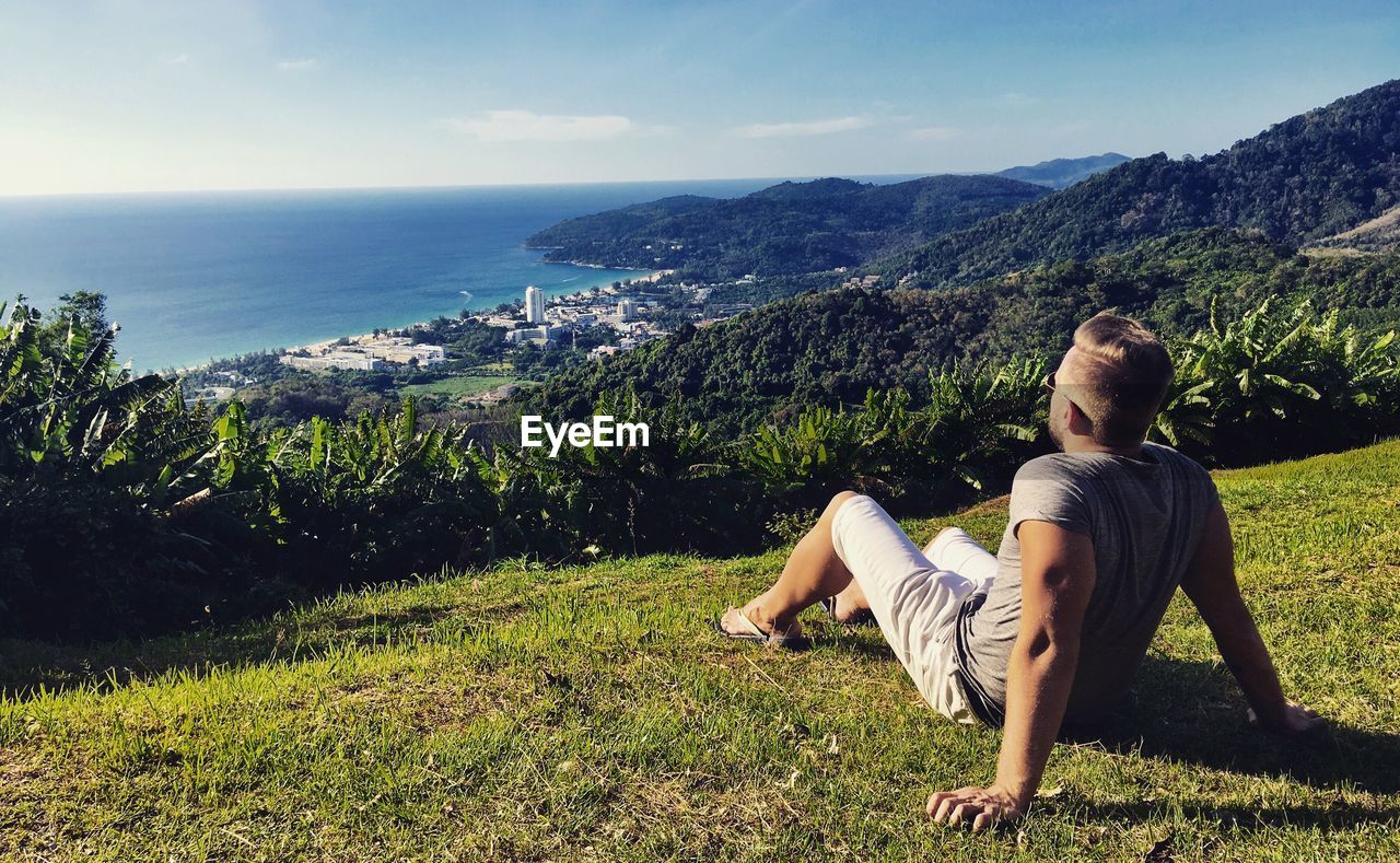 Young man sitting on grassy mountain against sea