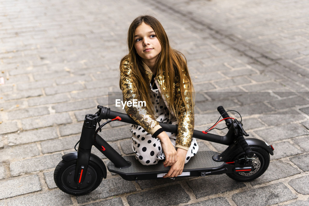 Portrait of girl wearing golden sequin jacket kneeling on folded e-scooter