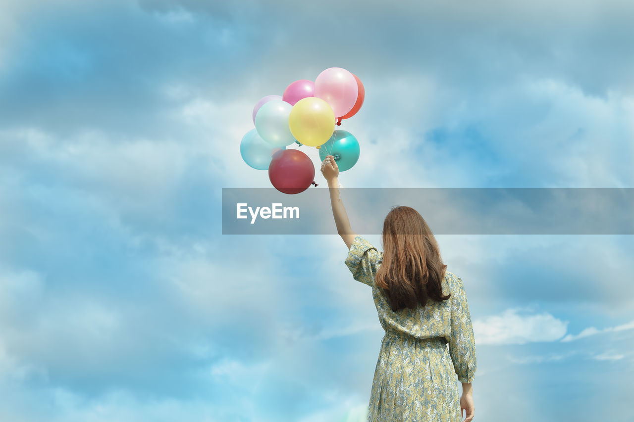 REAR VIEW OF WOMAN HOLDING BALLOONS WITH UMBRELLA AGAINST SKY