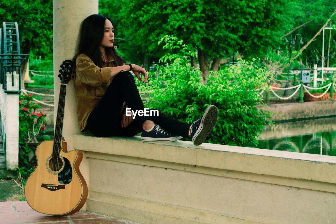 Full length of young woman sitting on retaining wall with guitar