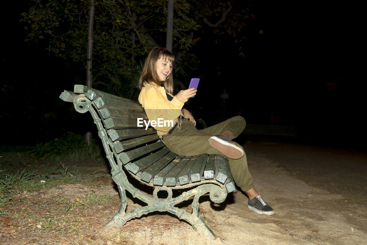 Full length of young woman using mobile phone sitting on bench at park