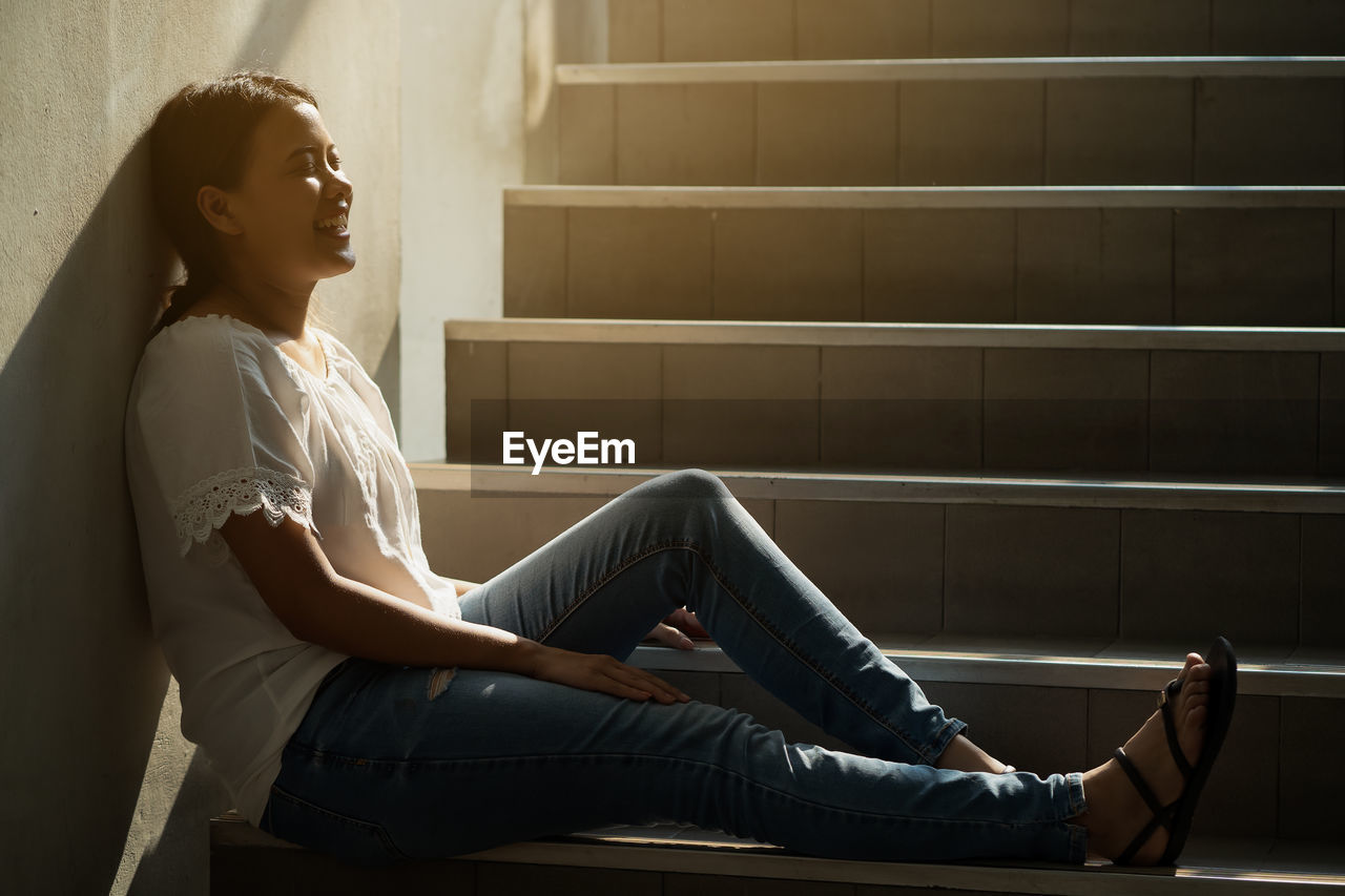 Side view of young woman sitting on staircase against wall