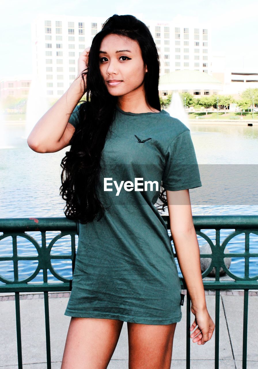 PORTRAIT OF BEAUTIFUL YOUNG WOMAN STANDING BY RAILING AGAINST WATER