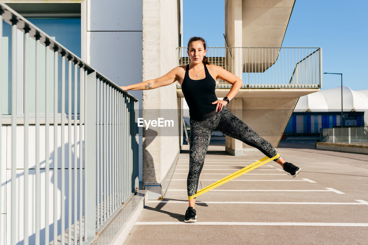 Slim female athlete in sportswear doing exercises with elastic band while standing near metal fence during workout in city and looking at camera resist