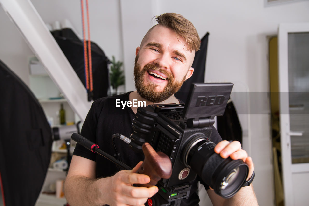 adult, occupation, technology, men, one person, camera, smiling, portrait, person, indoors, front view, happiness, beard, working, expertise, holding, facial hair, skill, filmmaking, professional occupation, young adult, arts culture and entertainment, equipment, digital slr, emotion