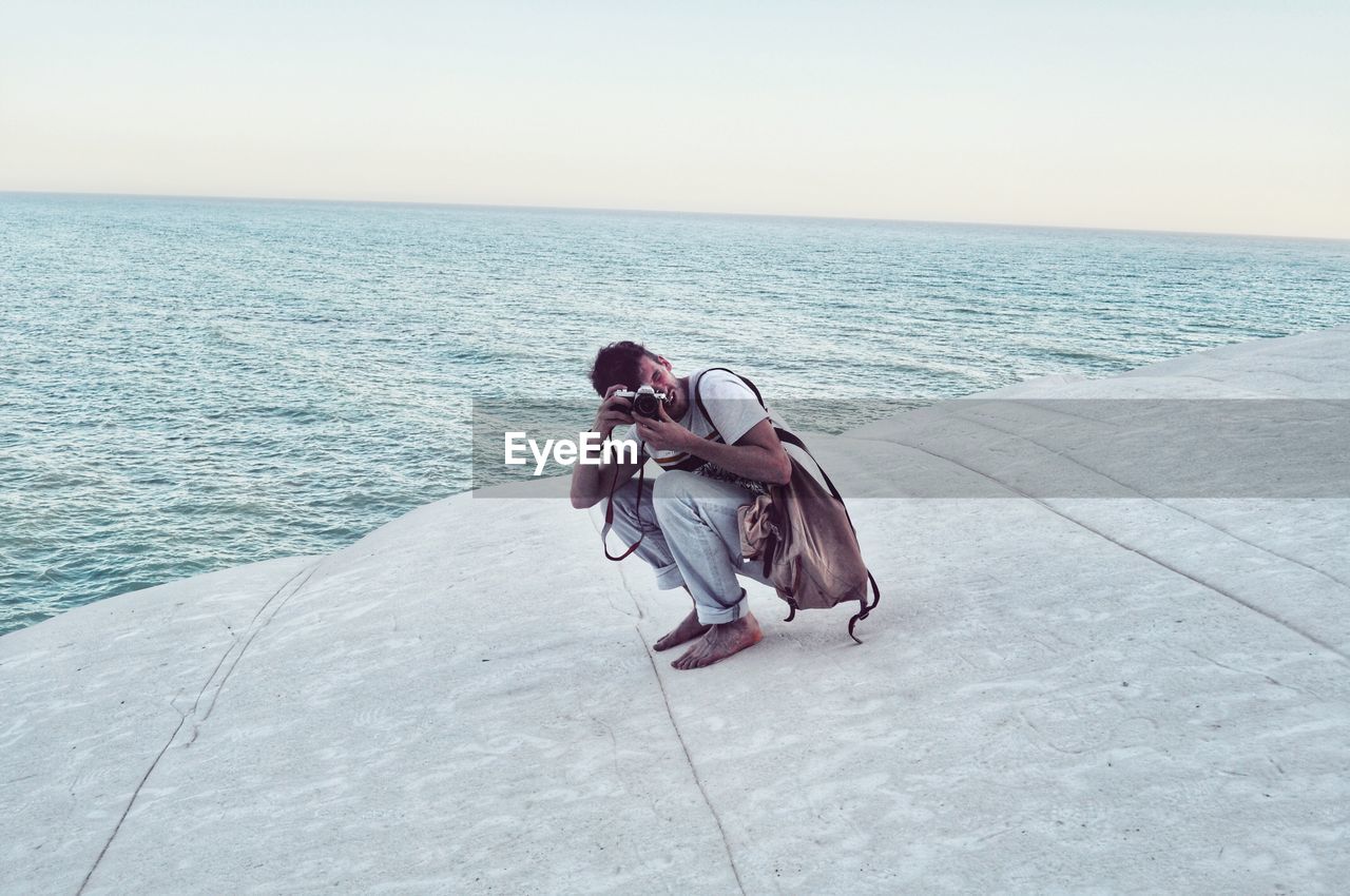 MAN SITTING ON SHORE AGAINST SKY