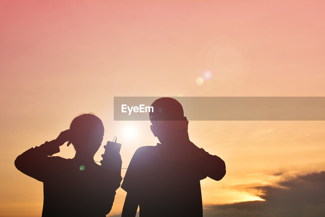 Silhouette girl with brother listening to music against sky during sunset