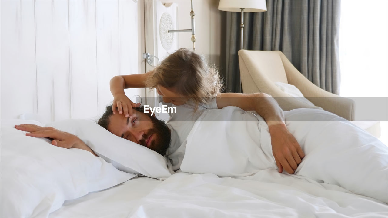 Cute girl opening eye of father lying on bed at home