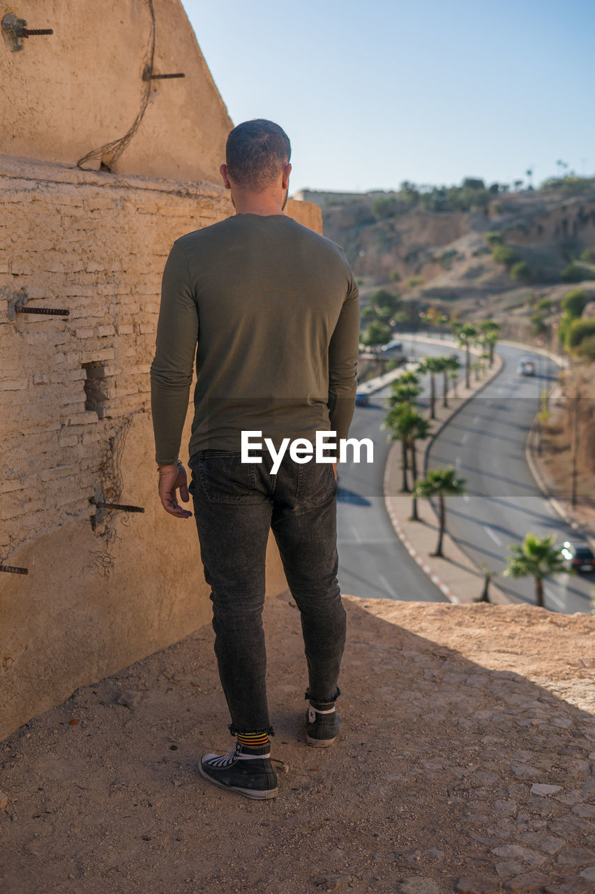 one person, men, full length, adult, rear view, architecture, nature, standing, day, casual clothing, travel, wall, sky, trousers, outdoors, clothing, lifestyles, sand, person, leisure activity, travel destinations, city, built structure, sunlight, walking, looking, history, road