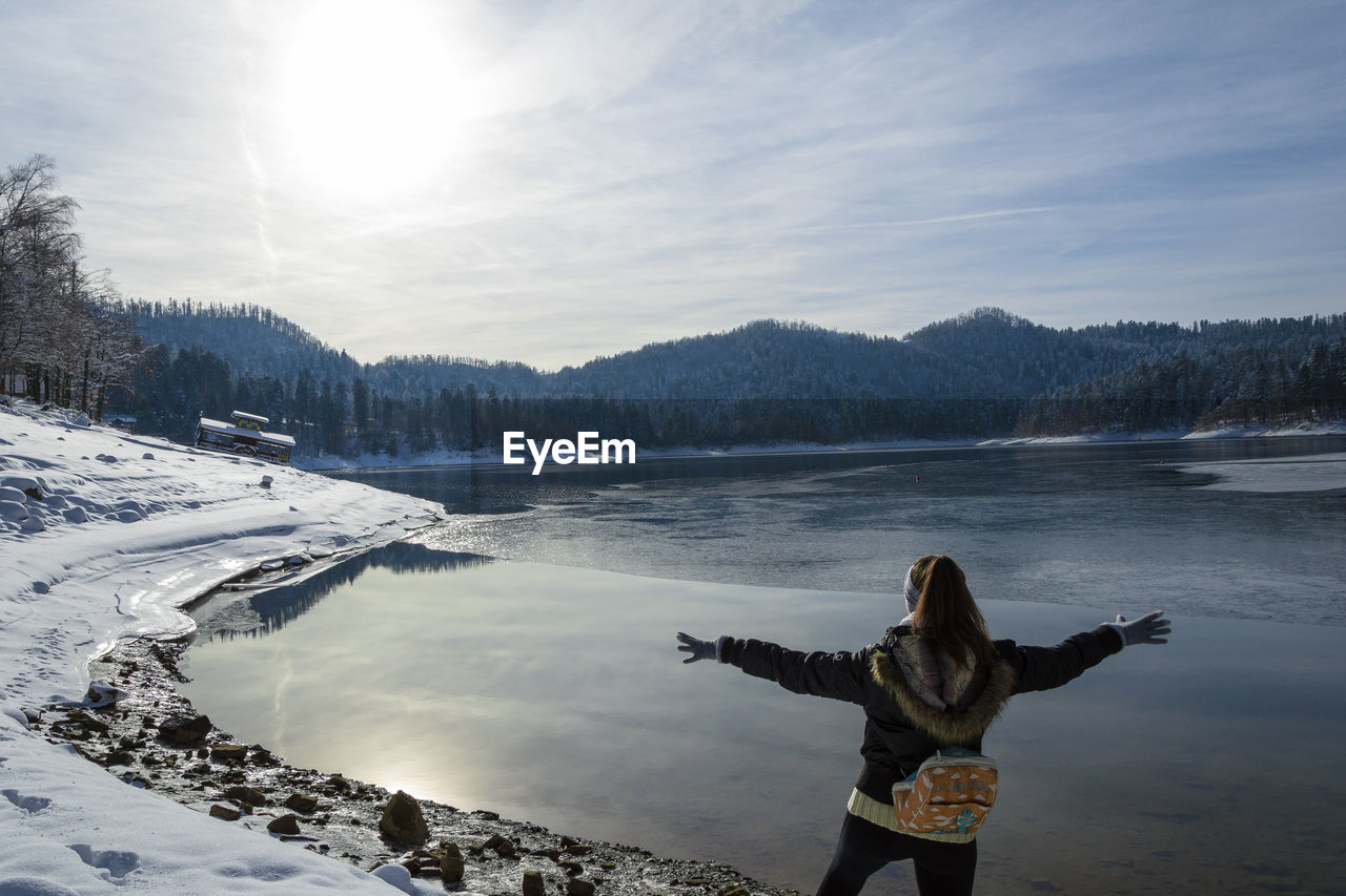 Rear view of woman with arms outstretched standing by lake against sky