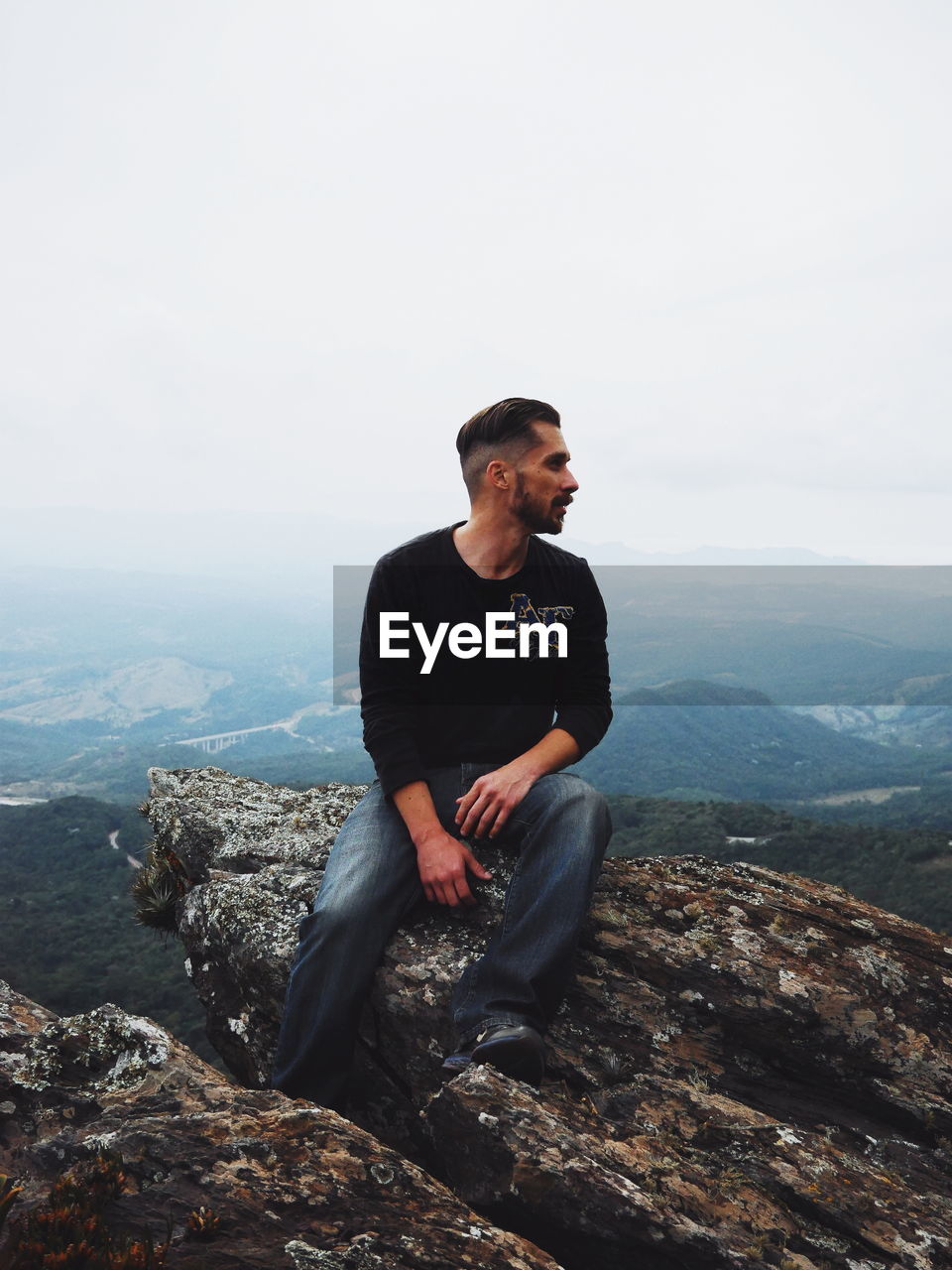 YOUNG MAN SITTING ON ROCK LOOKING AT MOUNTAIN