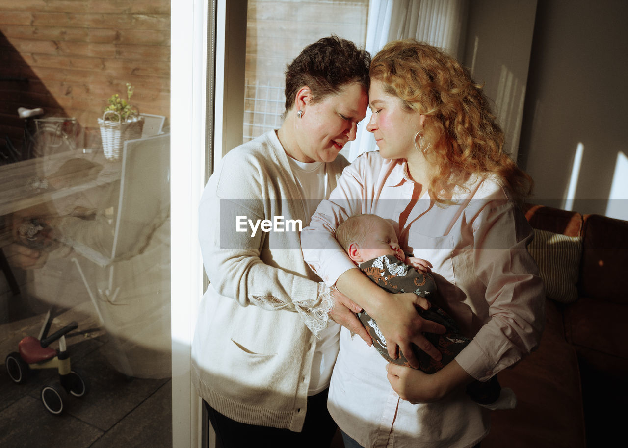 Side view of mother and daughter standing in front of window at home and holding a newborn son