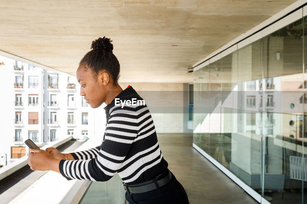 Side view of focused african american female in casual wear text messaging on cellphone while standing on balcony with glass wall