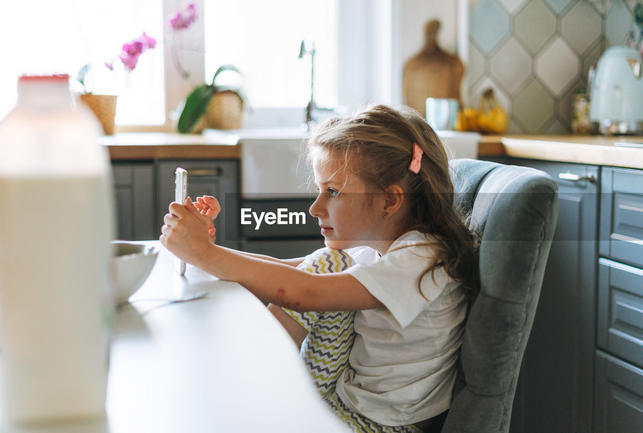Little girl with long hair in home clothes using mobile at dinning table in kitchen at home