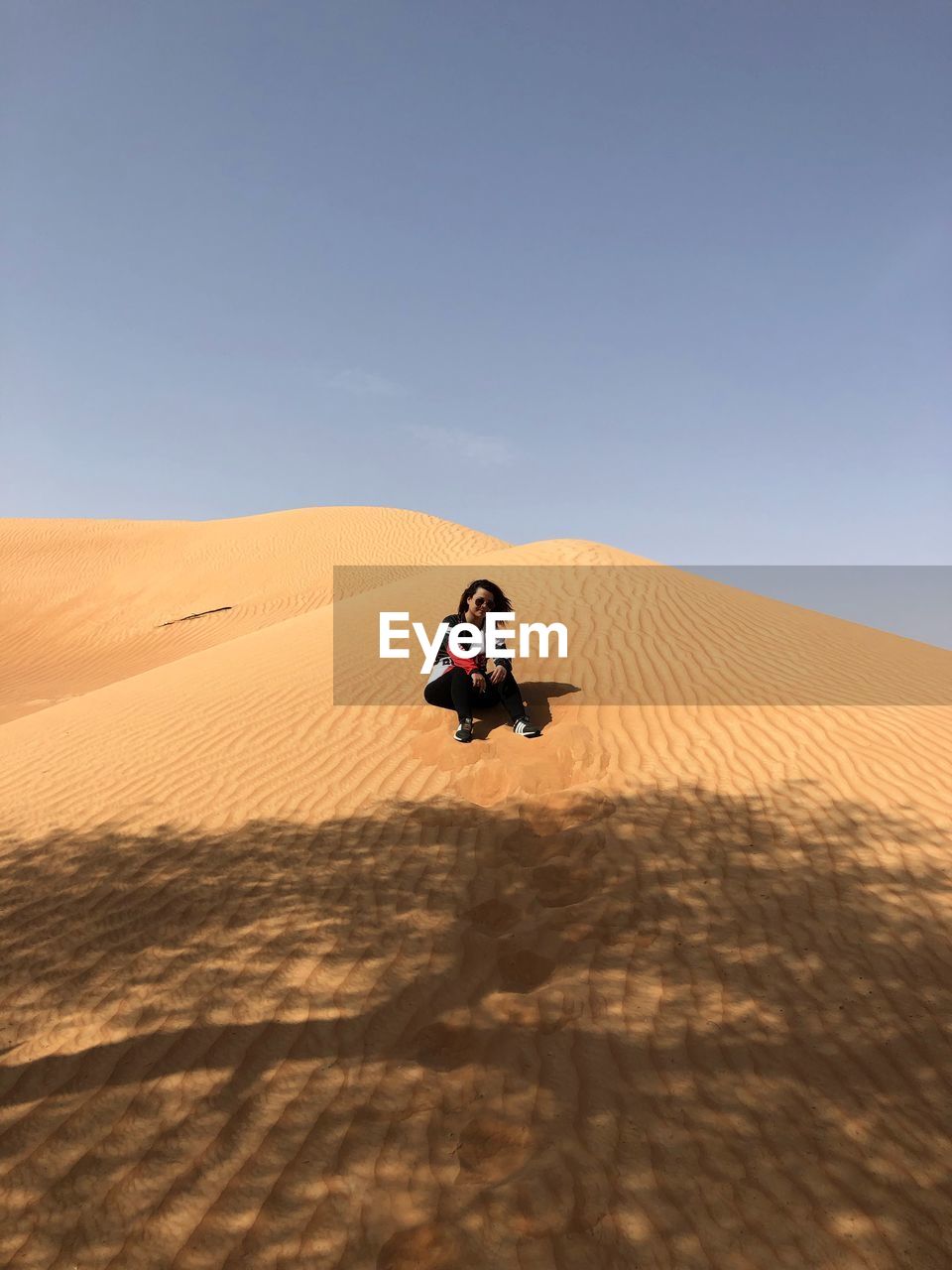 Low angle view of woman sitting on sand dune at desert against blue sky