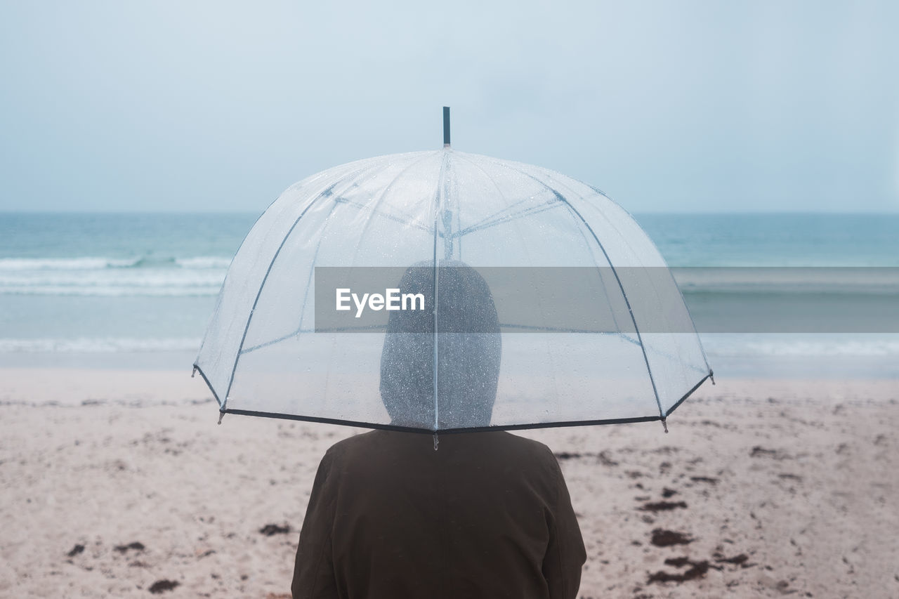 Back view of unrecognizable traveler in raincoat with transparent umbrella walking on empty sandy beach towards waving sea in cloudy day