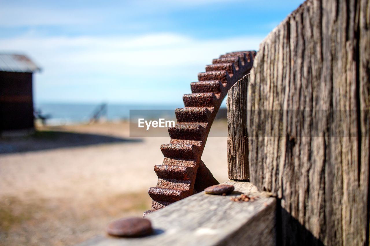 Close-up of rusty gear on wooden post at beach against sky