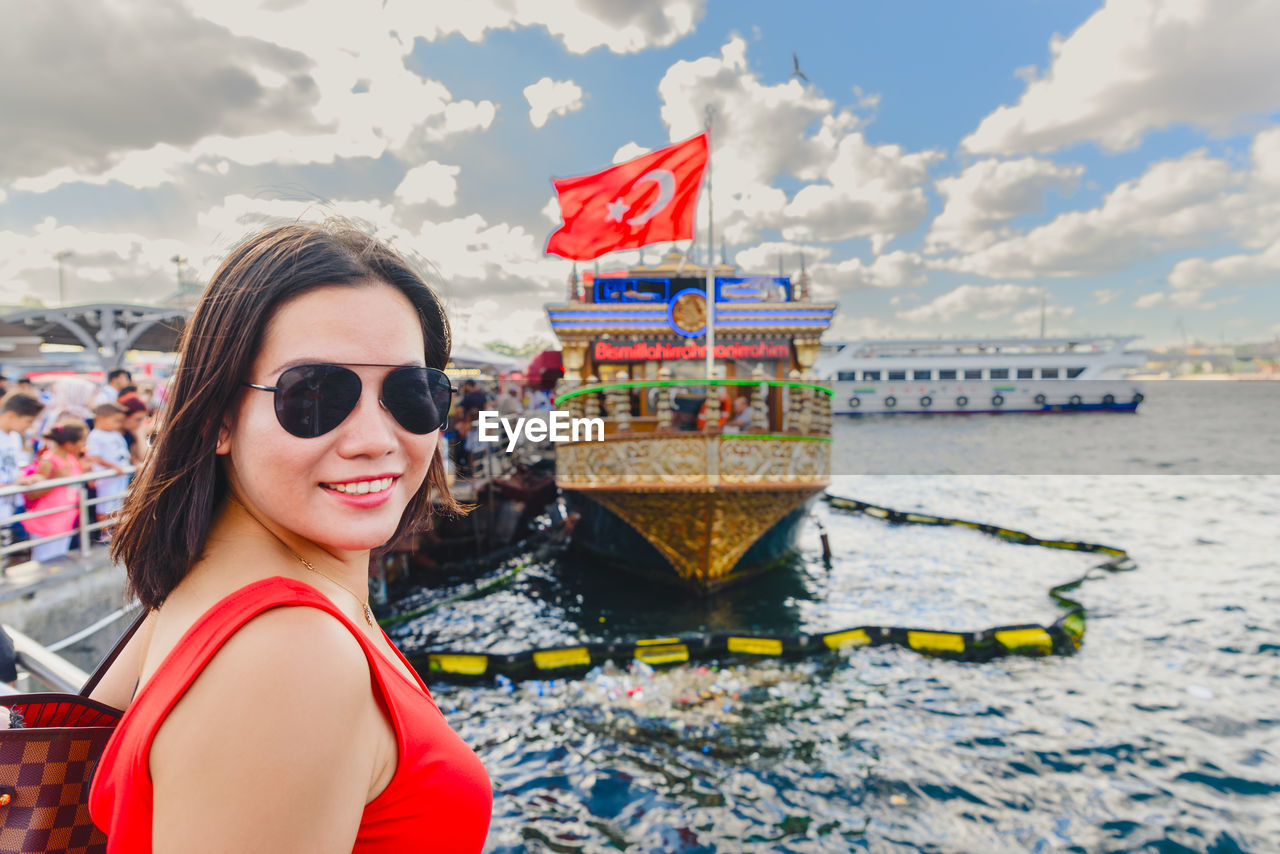 Portrait of woman wearing sunglasses pointing while standing by railing against sea