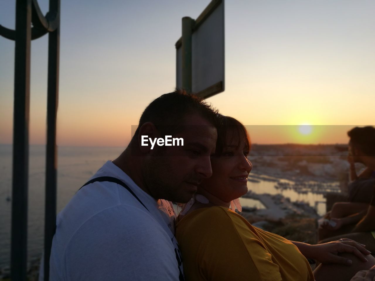 Couple looking away while standing by railing against sky during sunset