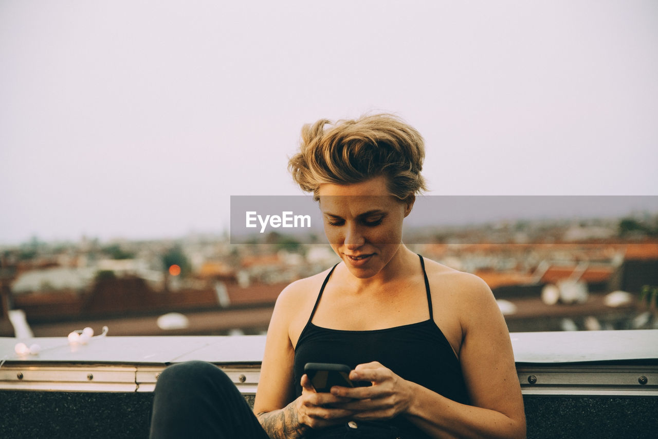 Smiling woman with short hair text messaging on smart phone while sitting on terrace against sky