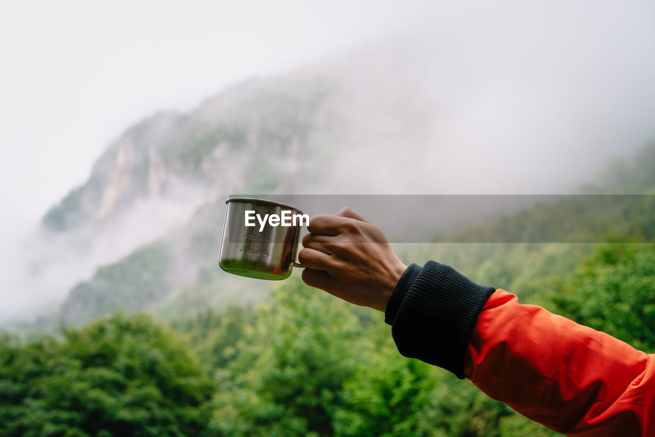 Midsection of person holding drinking cup against foggy mountains
