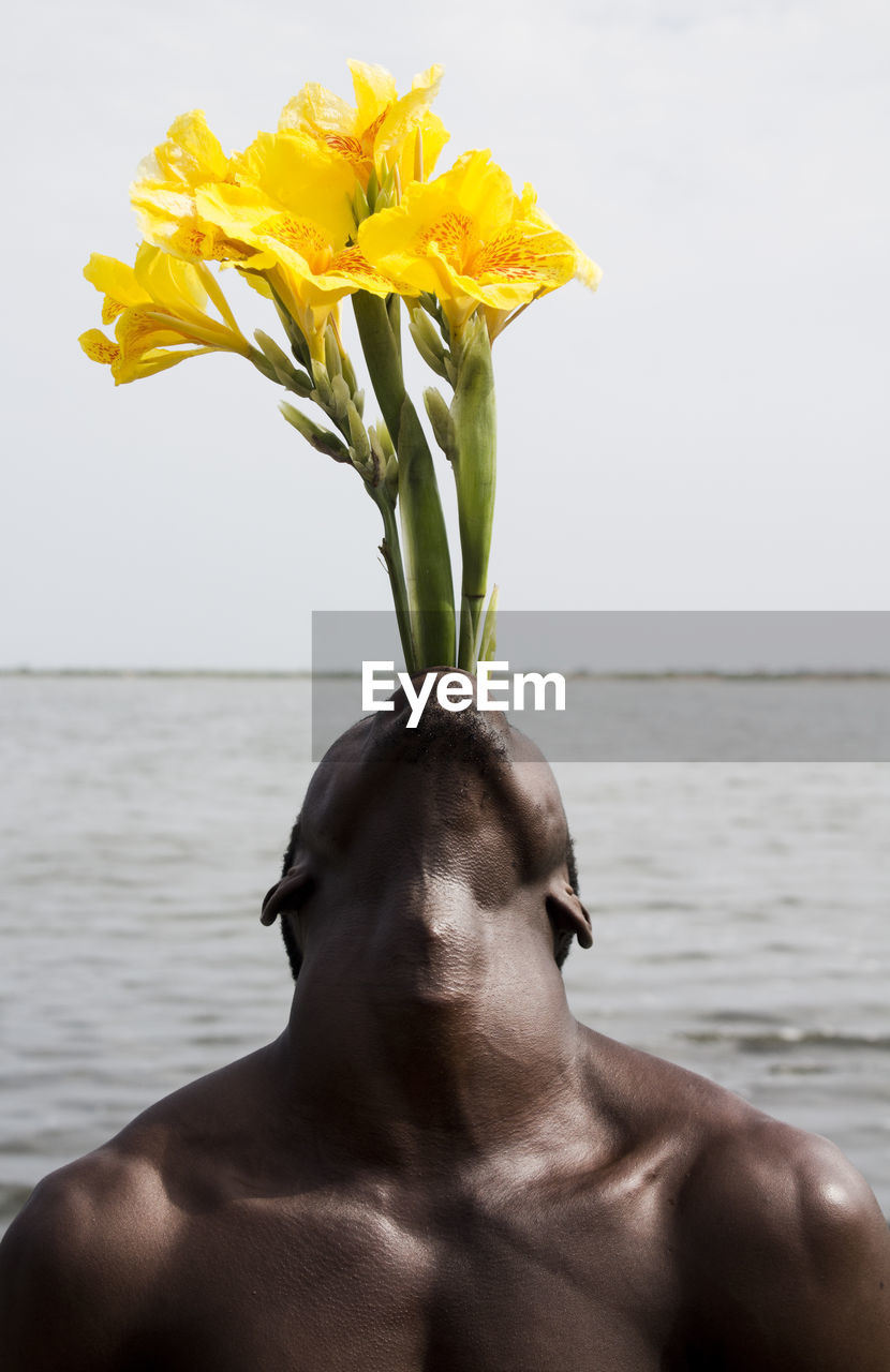 Close-up of shirtless man with flowers in mouth against sea