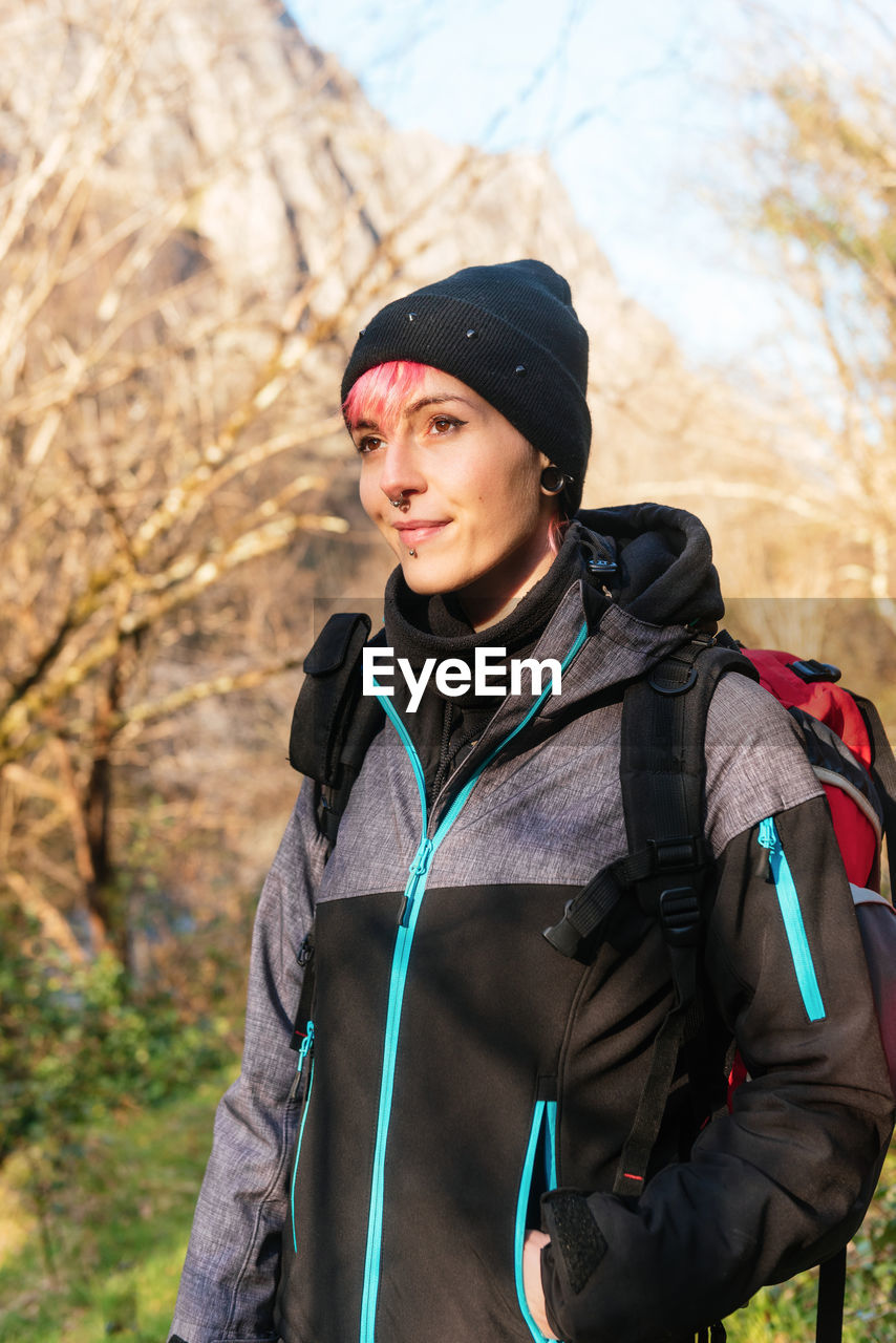 Positive female explorer wearing hat and outerwear with backpack looking away while standing blurred background during trekking in nature