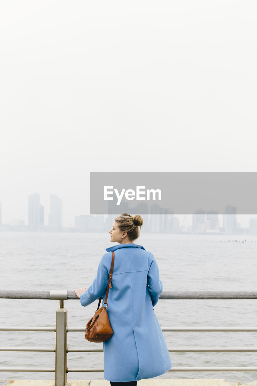 Young woman looking at view while standing by railing and river in city against clear sky